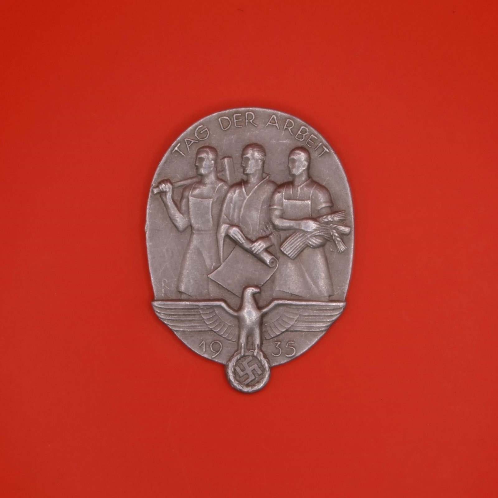 1935 "Tag der Arbeit" Labor Badge: An oval 1935 labor-day badge featuring three standing figures-a worker with a hammer, a man holding a rolled document, and a woman with a bundle of wheat. Below them is an eagle above the date "1935"