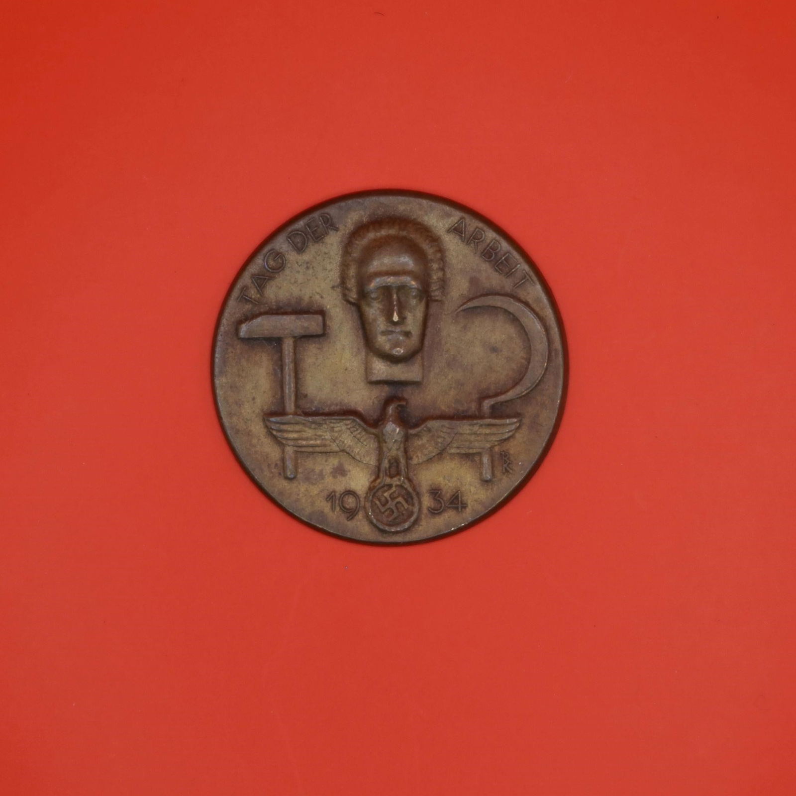 1934 German Labor Day Badge (Bronze Tone): A round 1934 commemorative German Labor Day badge in bronze tone. The front shows a hammer, a sickle, and a stylized face above an eagle emblem. The back has the original pin, though aged. Displays we