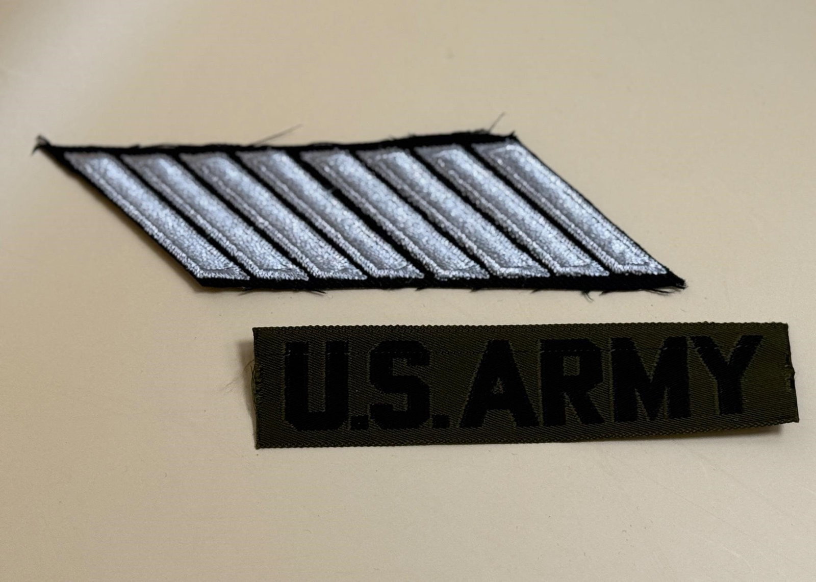 U.S. Army Uniform Patch & Name Tag: The top patch with silver stripes on a black background is likely a rank or service stripe patch (hashmark). Each stripe typically represents a period of service, often three years.The bottom patch