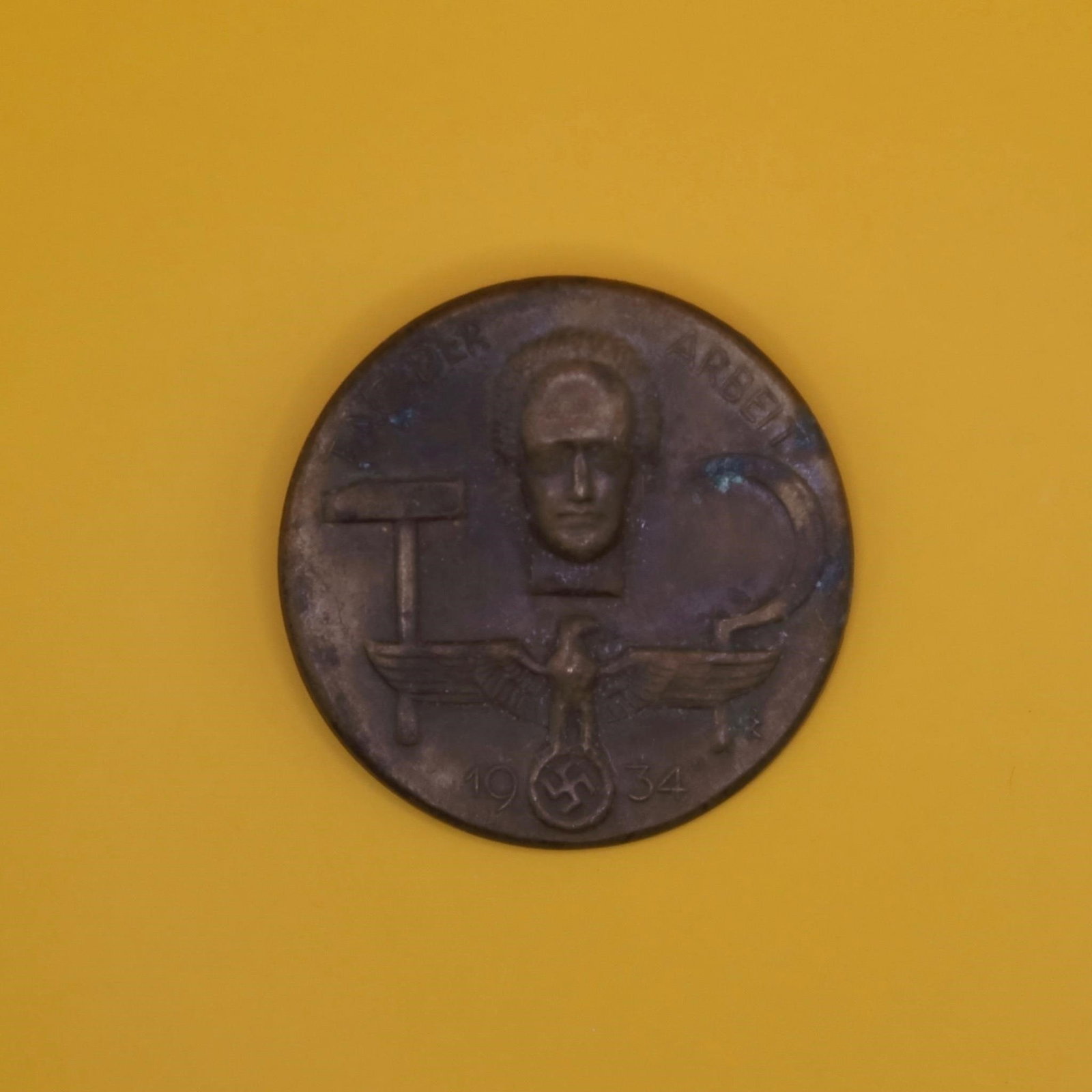 1934 German Labor Day Badge (Bronze Tone): A round 1934 commemorative German Labor Day badge in bronze tone. The front shows a hammer, a sickle, and a stylized face above an eagle emblem. The back has the original pin, though aged. Displays