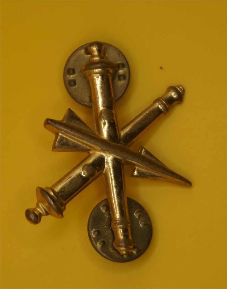 US Army Air Defense Artillery Branch Insignia: It features a missile surmounting two crossed field guns, all in gold-colored metal. The insignia has been in use since 1957, with the Air Defense Artillery established as a basic branch in 1968. It