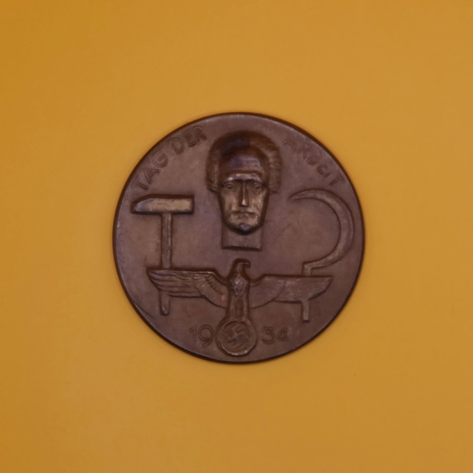 1934 German Labor Day Badge (Bronze Tone): A round 1934 commemorative German Labor Day badge in bronze tone. The front shows a hammer, a sickle, and a stylized face above an eagle emblem. Displays wear and patina consistent with age.