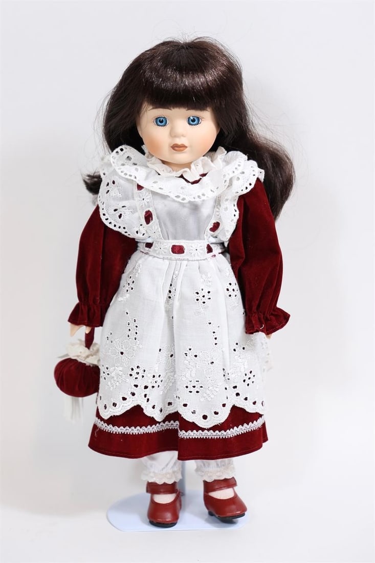 Porcelain Doll in Red Velvet Dress with Lace Apron: Charming porcelain doll featuring a finely detailed face with hand-painted features, blue eyes, and long dark brown hair. The doll is dressed in a rich red velvet outfit with a white lace apron,