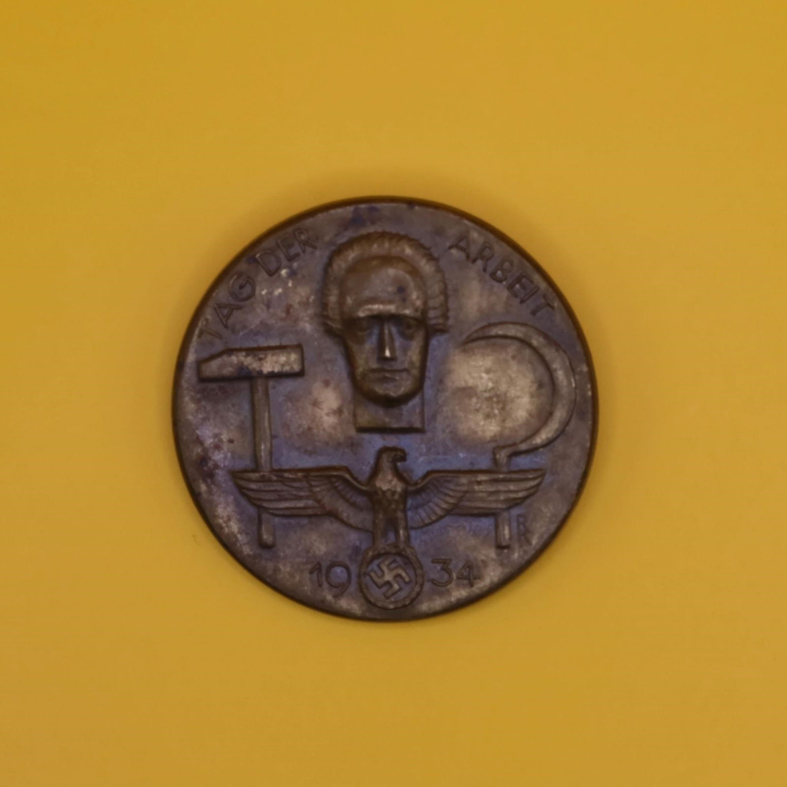 1934 German Labor Day Badge (Bronze Tone): A round 1934 commemorative German Labor Day badge in bronze tone. The front shows a hammer, a sickle, and a stylized face above an eagle emblem. The back has the original pin, though aged. Displays