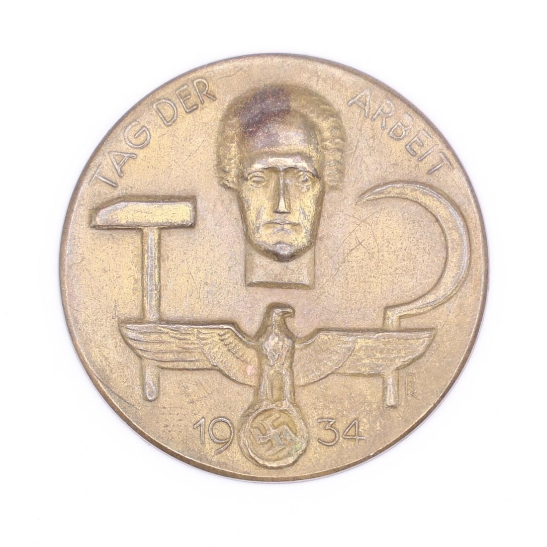 1934 German Labor Day Badge (Bronze Tone): A round 1934 commemorative German Labor Day badge in bronze tone. The front shows a hammer, a sickle, and a stylized face above an eagle emblem. Displays wear and patina consistent with age.