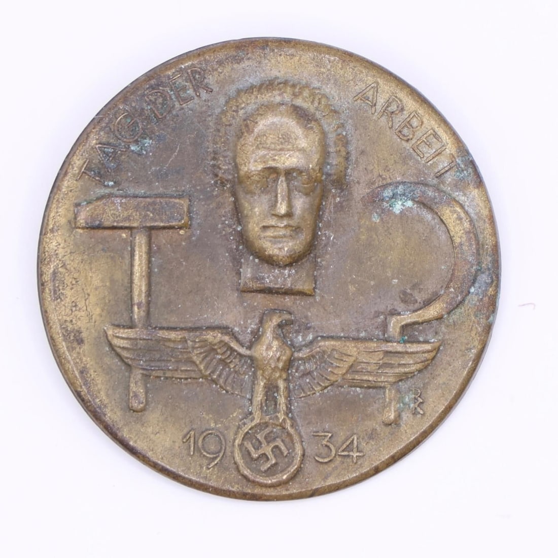 1934 German Labor Day Badge (Bronze Tone): A round 1934 commemorative German Labor Day badge in bronze tone. The front shows a hammer, a sickle, and a stylized face above an eagle emblem. The back has the original pin, though aged. Displays