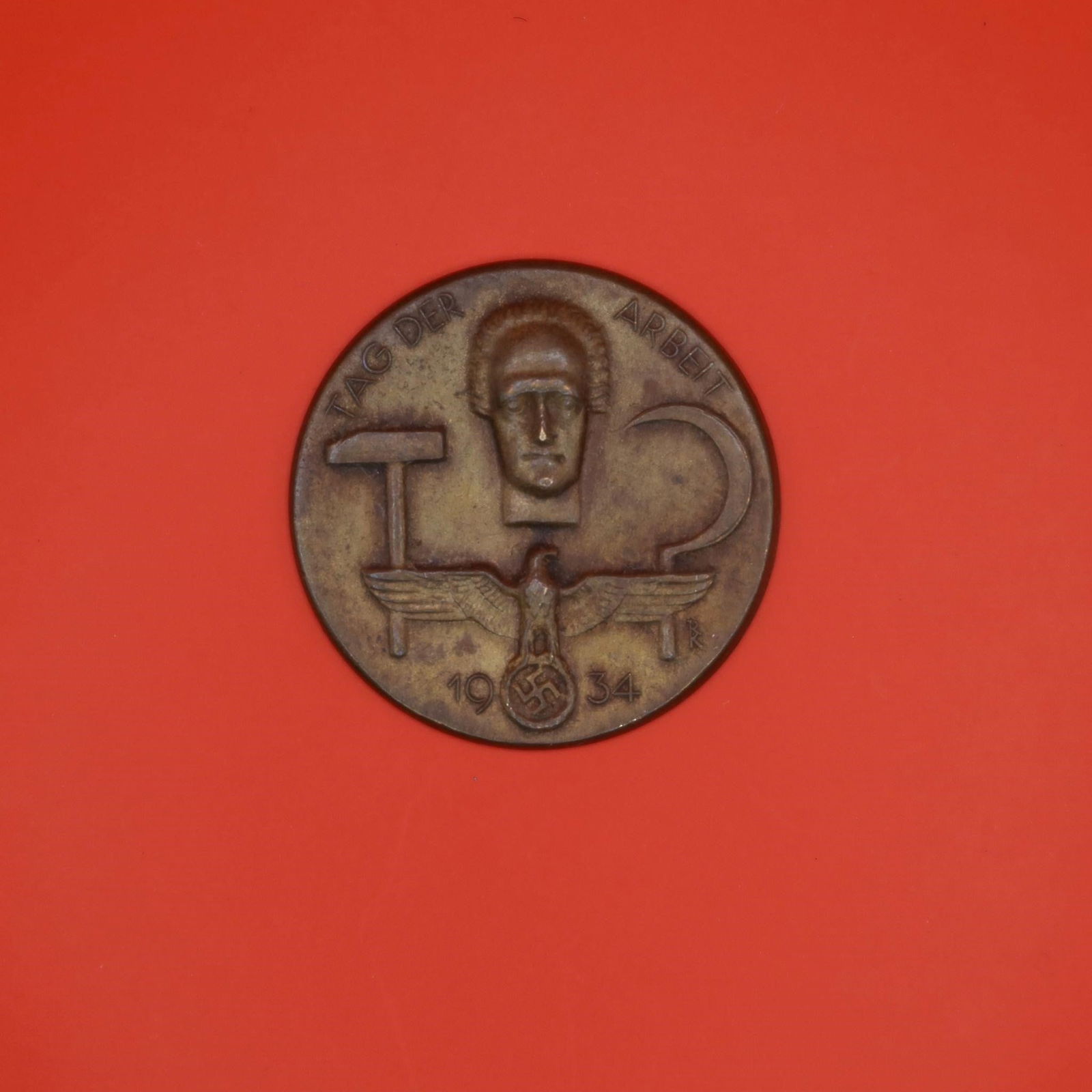 1934 German Labor Day Badge (Bronze Tone): A round 1934 commemorative German Labor Day badge in bronze tone. The front shows a hammer, a sickle, and a stylized face above an eagle emblem. The back has the original pin, though aged. Displays