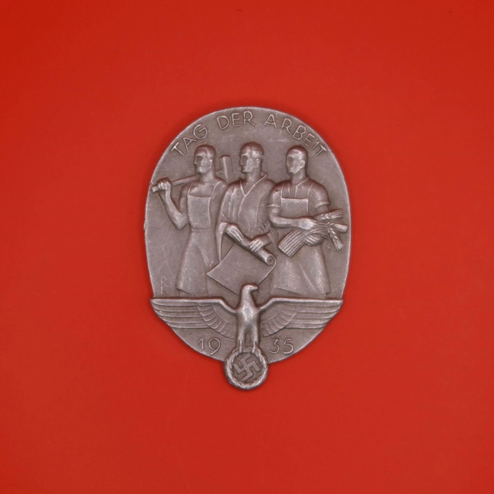 1935 "Tag der Arbeit" Labor Badge: An oval 1935 labor-day badge featuring three standing figures-a worker with a hammer, a man holding a rolled document, and a woman with a bundle of wheat. Below them is an eagle above the date "1935"
