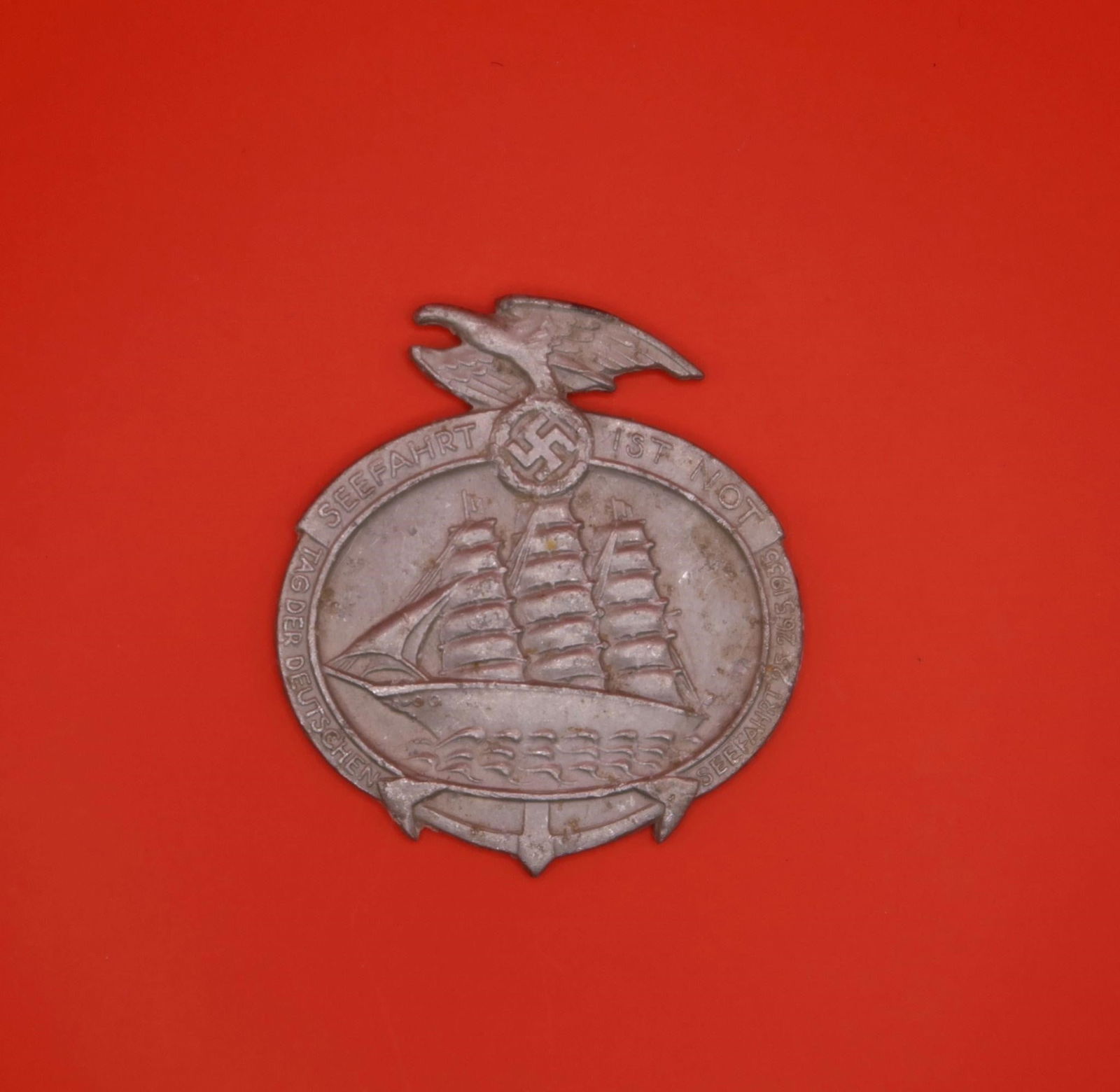 1935 "Seefahrt ist Not" Tinnie Badge: A lightweight metal event badge issued for the 1935 Tag der Deutschen Seefahrt (Day of German Seafaring). The front shows a multi-masted ship on the water, surrounded by the event wording, topped by