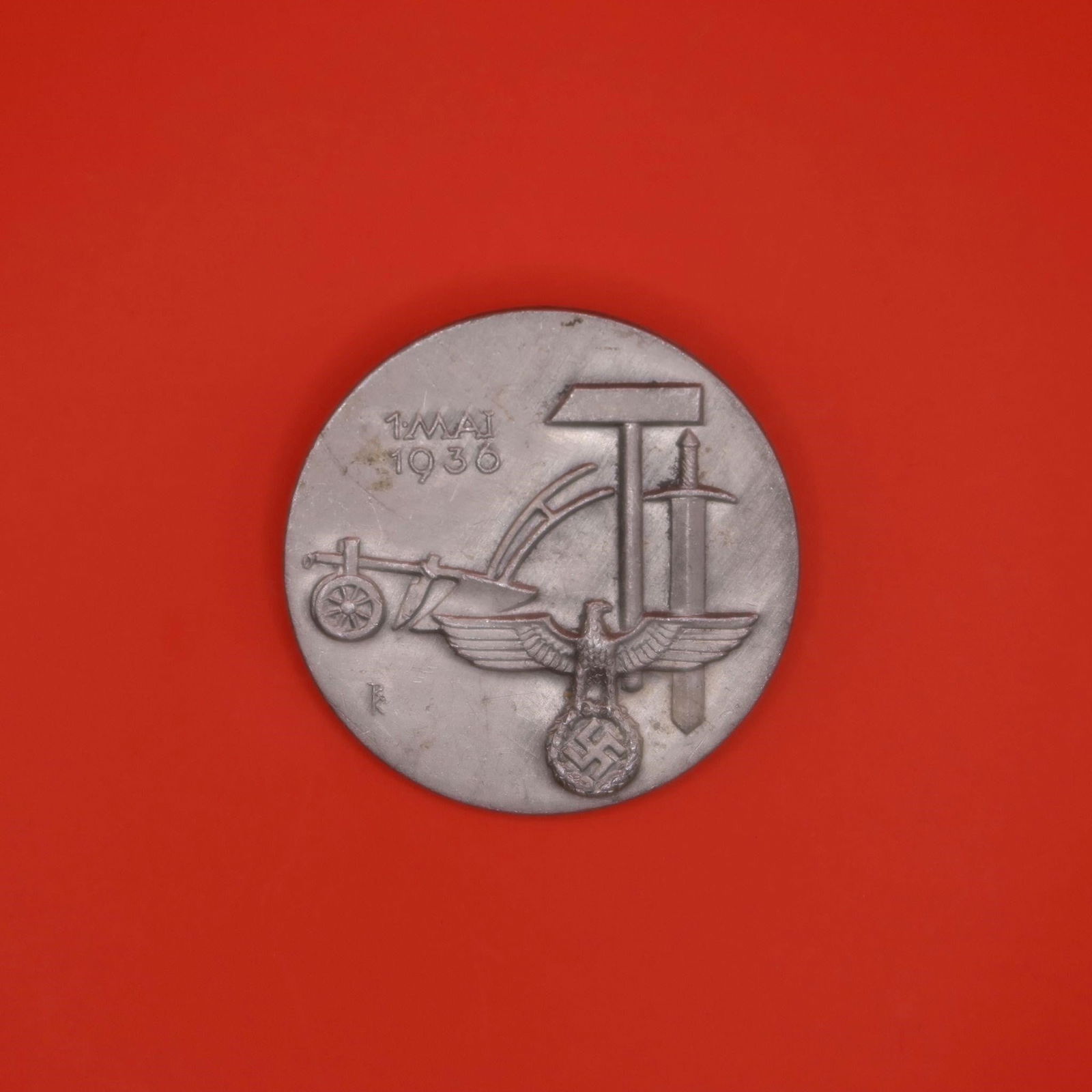 1936 Tag der Arbeit Tin Day Badge: This round 1936 German "Tag der Arbeit" badge is made of a light-colored metal, likely tin. The front shows a plow, hammer, and sword grouped together above an eagle emblem. The date "1. Mai 1936"