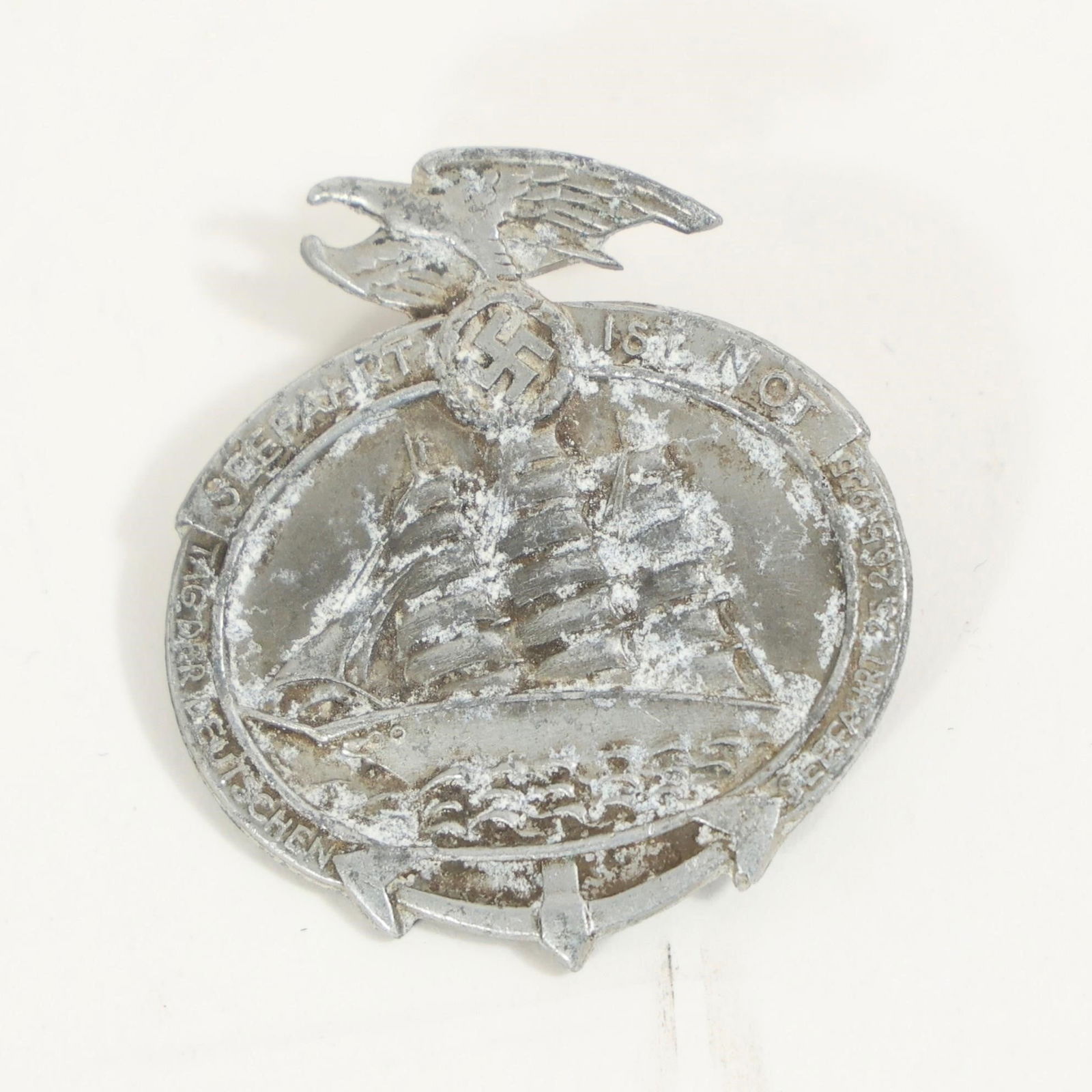 German National Sea Travel Day Badge: A lightweight metal event badge showing a three-masted sailing ship over waves, topped by an eagle clutching emblem. The front ring carries the event inscription, and the back has a simple pin