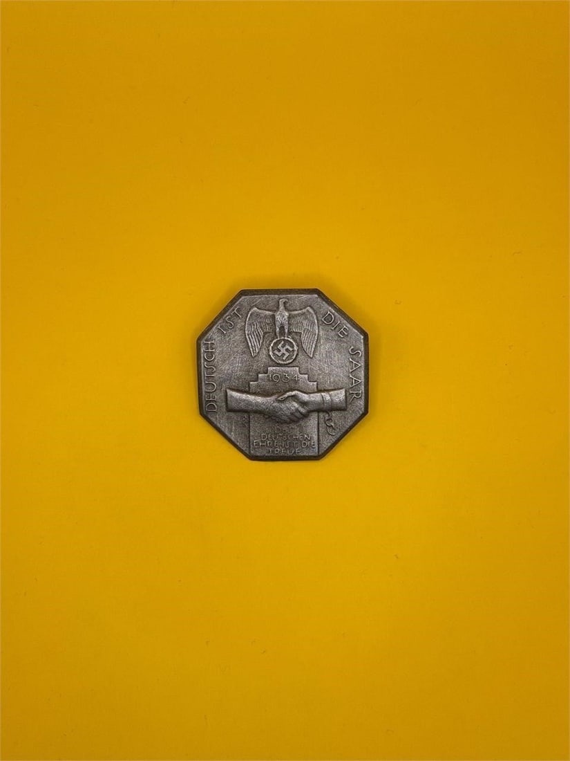 1934 NSDAP "Deutsch ist die Saar" Badge: Issued in 1934, this NSDAP badge commemorated the Saar region's return to Germany. It features an eagle clutching a wreathed swastika above two shaking hands, symbolizing unity and loyalty, and was