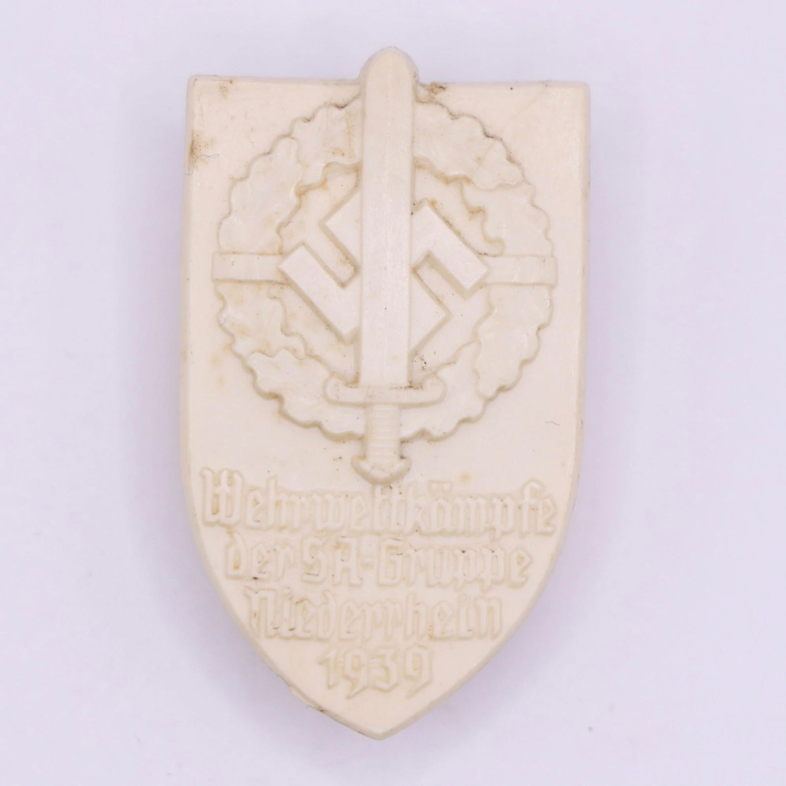 1939 SA Group Niederrhein Sport Day Badge: White protoplastic badge, approximately 35 × 50 mm, originally with bronze-colored paint that has faded over time. Features a sword with SA insignia on the pommel over an oak leaf wreath, with