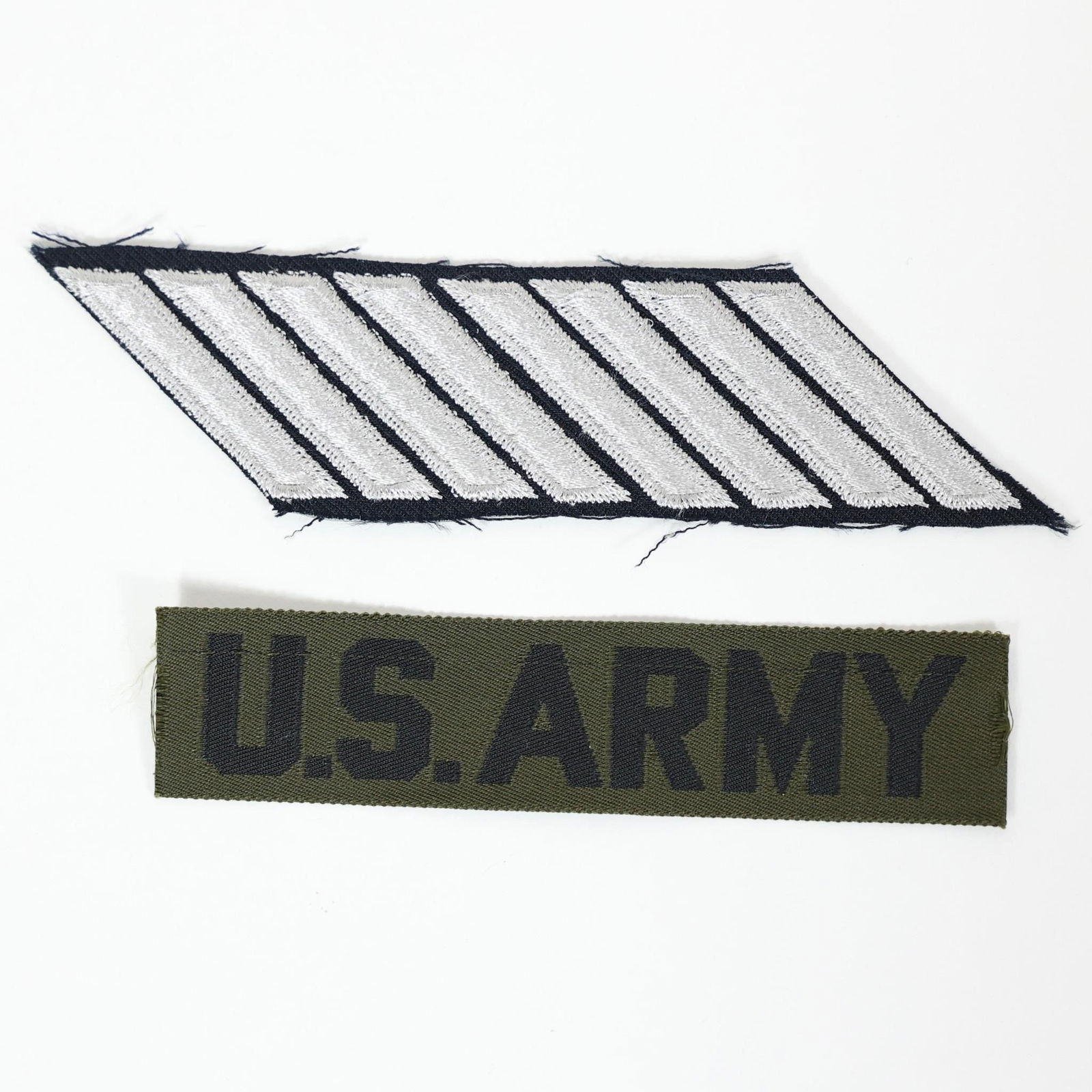 U.S. Army Uniform Patch & Name Tag: The top patch with silver stripes on a black background is likely a rank or service stripe patch (hashmark). Each stripe typically represents a period of service, often three years.The bottom patch is
