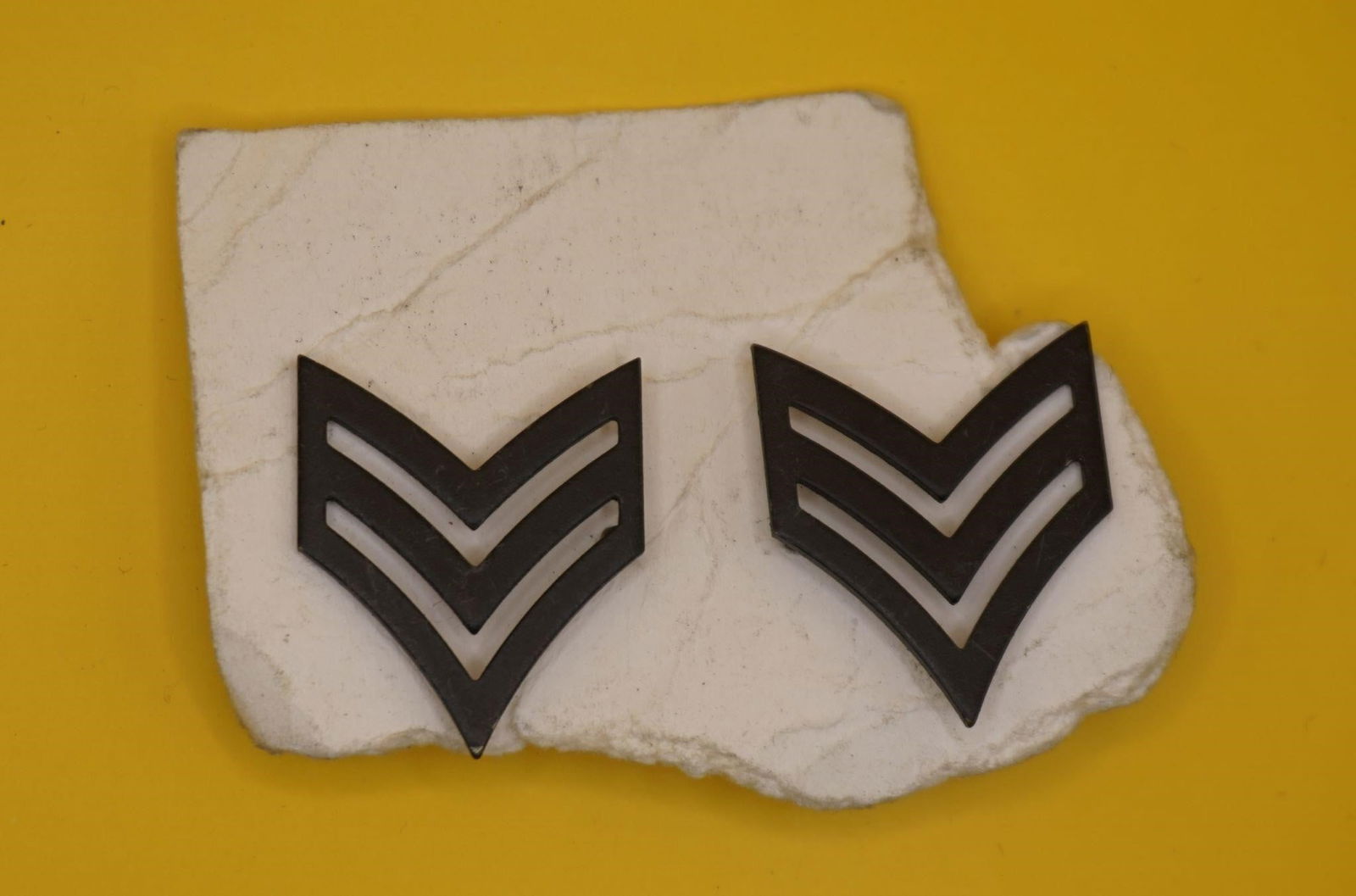 WWII US Army Sergeant Rank Insignia Pins: This is a pair of U.S. Army Sergeant rank insignia pins. The insignia features three chevrons, indicating the rank of Sergeant (E-5). The pins are a black metal, subdued design. They are authorized