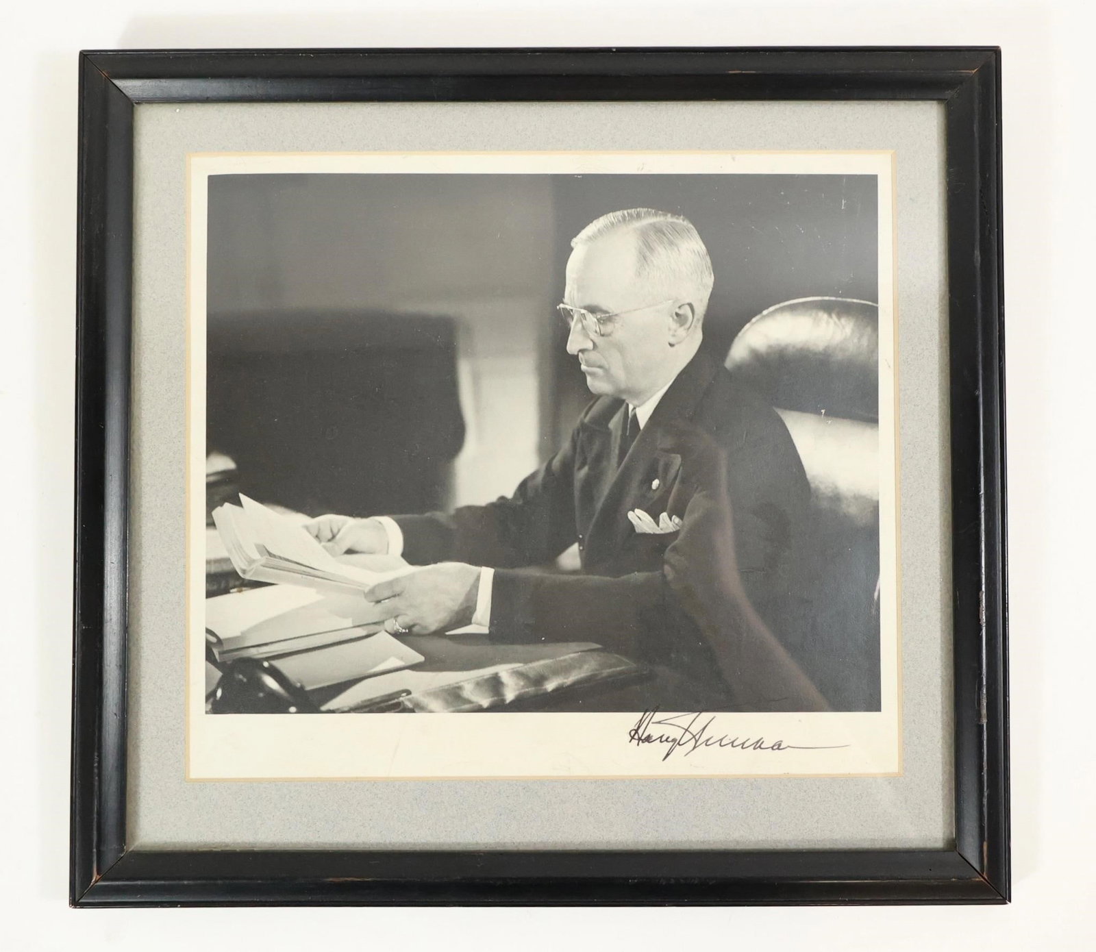 Harry Truman Signed Photo: Harry Truman black-and-white photograph showing him seated at a desk reading papers, with his original signature visible on the lower right of the image. Presented in a black frame with light matting.