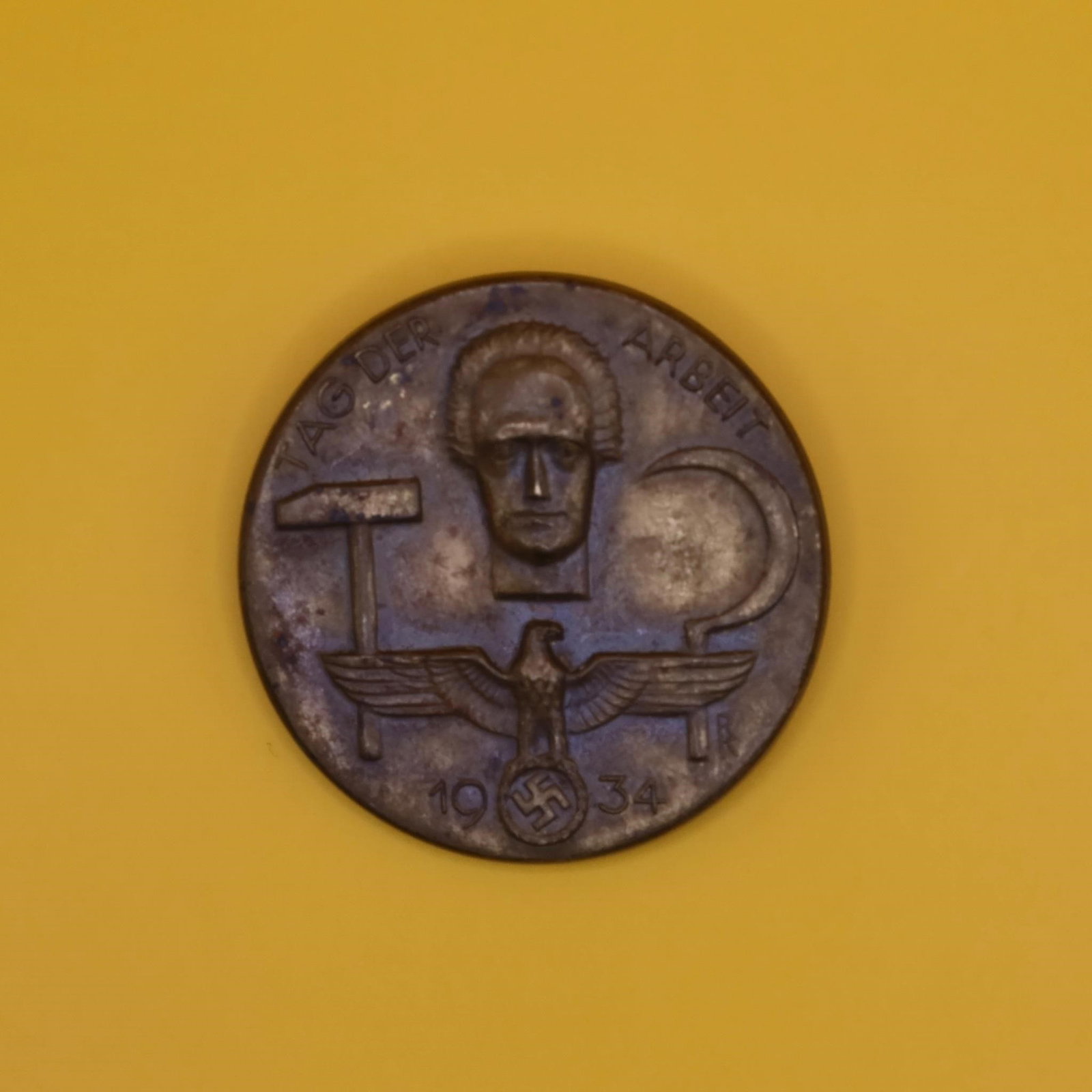 1934 German Labor Day Badge (Bronze Tone): A round 1934 commemorative German Labor Day badge in bronze tone. The front shows a hammer, a sickle, and a stylized face above an eagle emblem. The back has the original pin, though aged. Displays