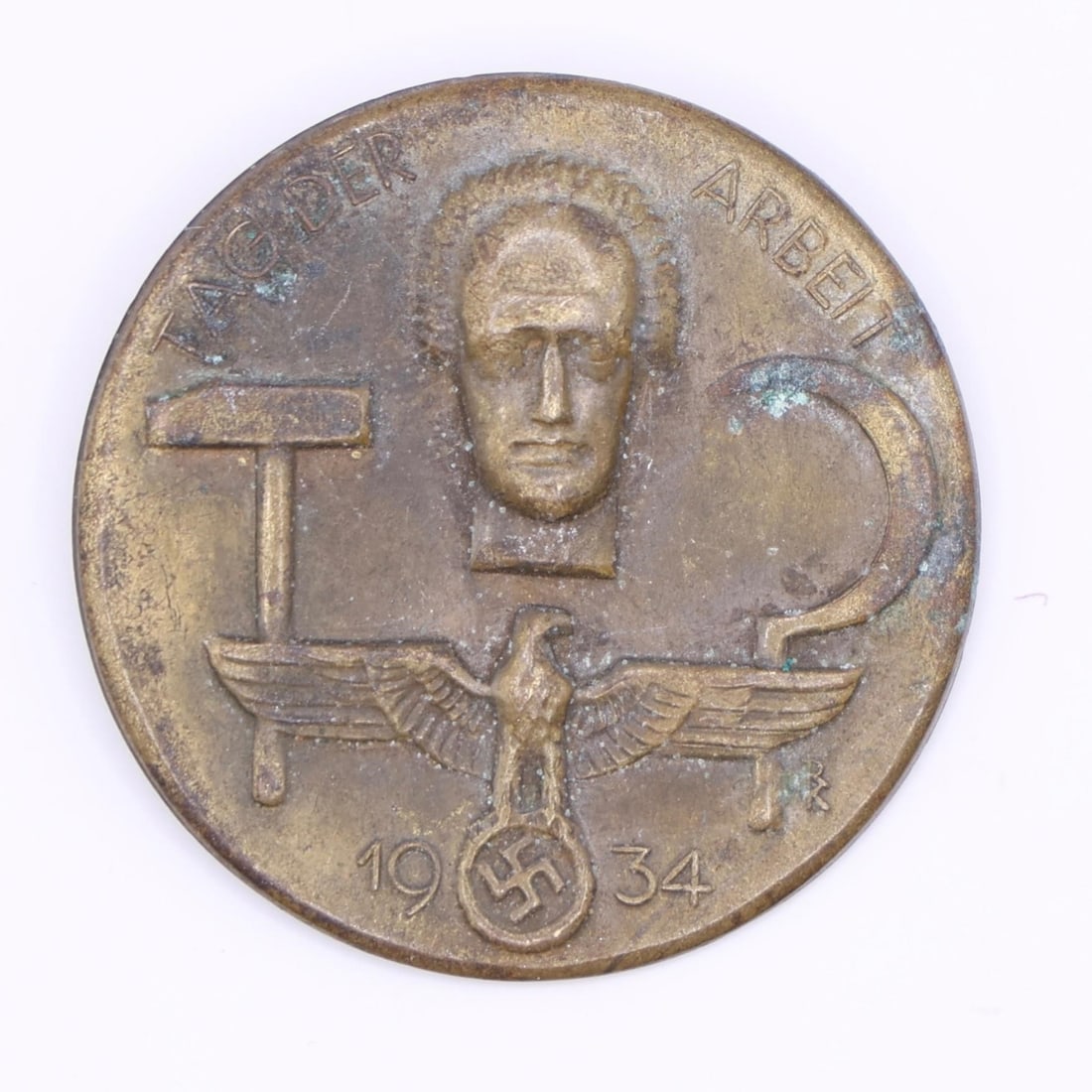 1934 German Labor Day Badge (Bronze Tone): A round 1934 commemorative German Labor Day badge in bronze tone. The front shows a hammer, a sickle, and a stylized face above an eagle emblem. The back has the original pin, though aged. Displays