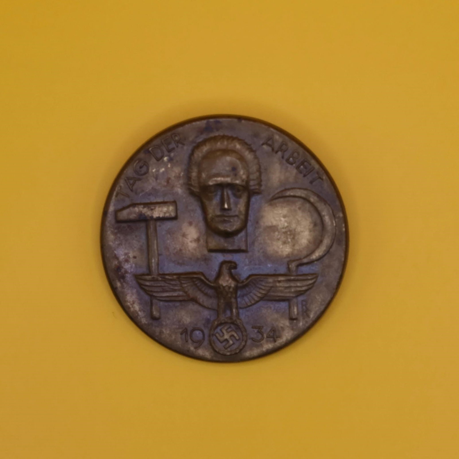 1934 German Labor Day Badge (Bronze Tone): A round 1934 commemorative German Labor Day badge in bronze tone. The front shows a hammer, a sickle, and a stylized face above an eagle emblem. The back has the original pin, though aged. Displays