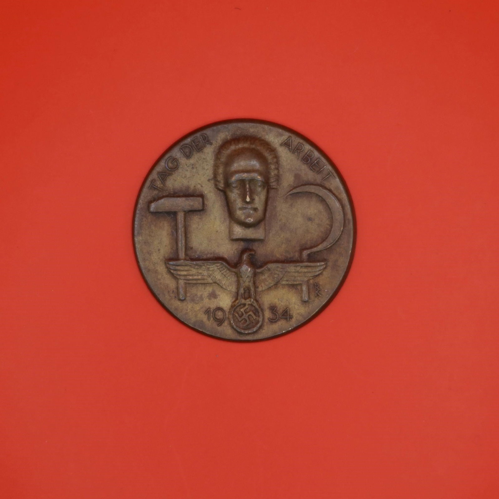 1934 German Labor Day Badge (Bronze Tone): A round 1934 commemorative German Labor Day badge in bronze tone. The front shows a hammer, a sickle, and a stylized face above an eagle emblem. The back has the original pin, though aged. Displays