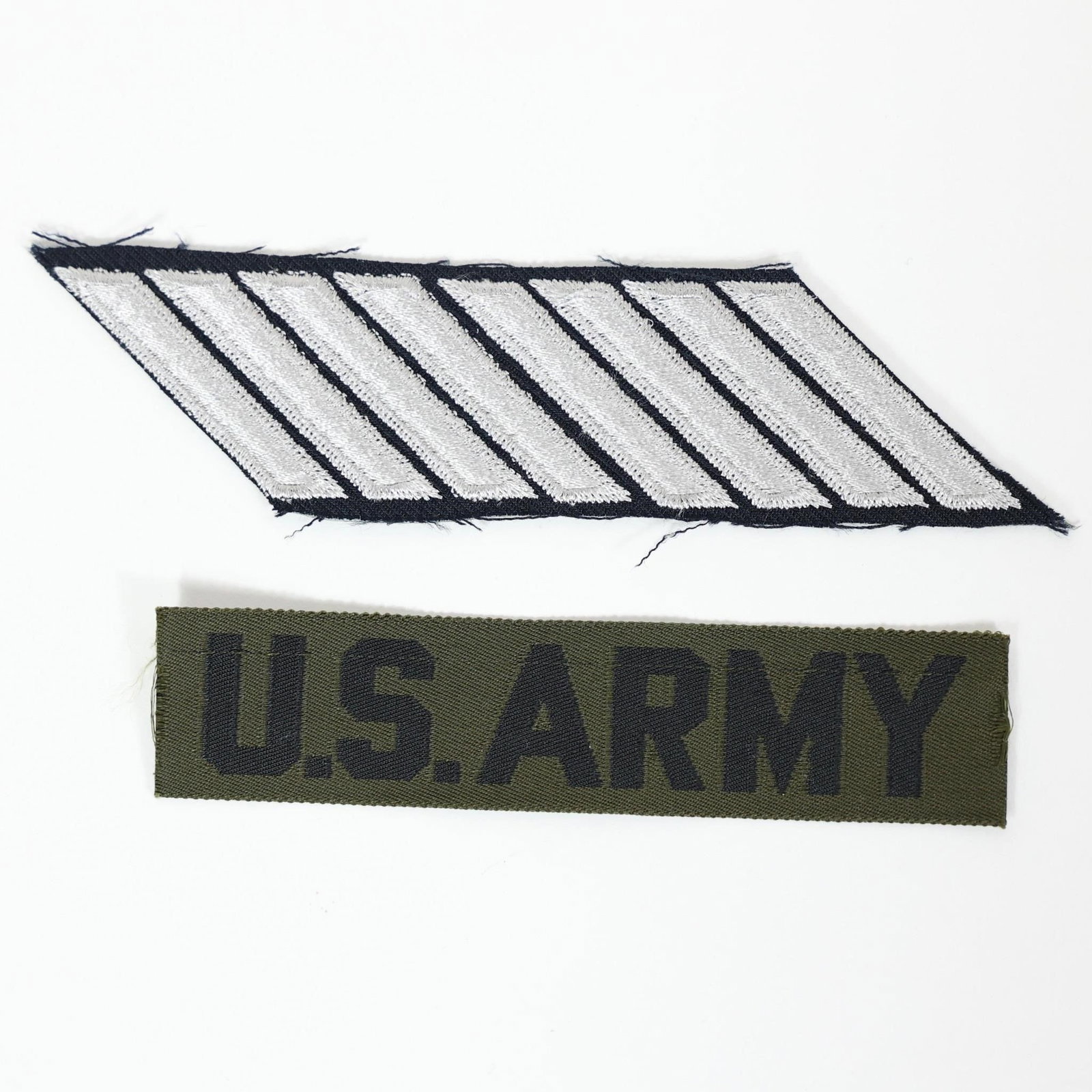 U.S. Army Uniform Patch & Name Tag: The top patch with silver stripes on a black background is likely a rank or service stripe patch (hashmark). Each stripe typically represents a period of service, often three years.The bottom patch