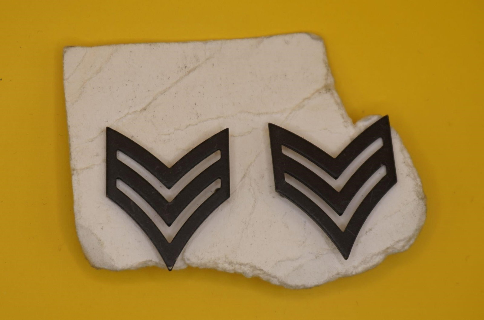 WWII US Army Sergeant Rank Insignia Pins: This is a pair of U.S. Army Sergeant rank insignia pins. The insignia features three chevrons, indicating the rank of Sergeant (E-5). The pins are a black metal, subdued design. They are authorized fo