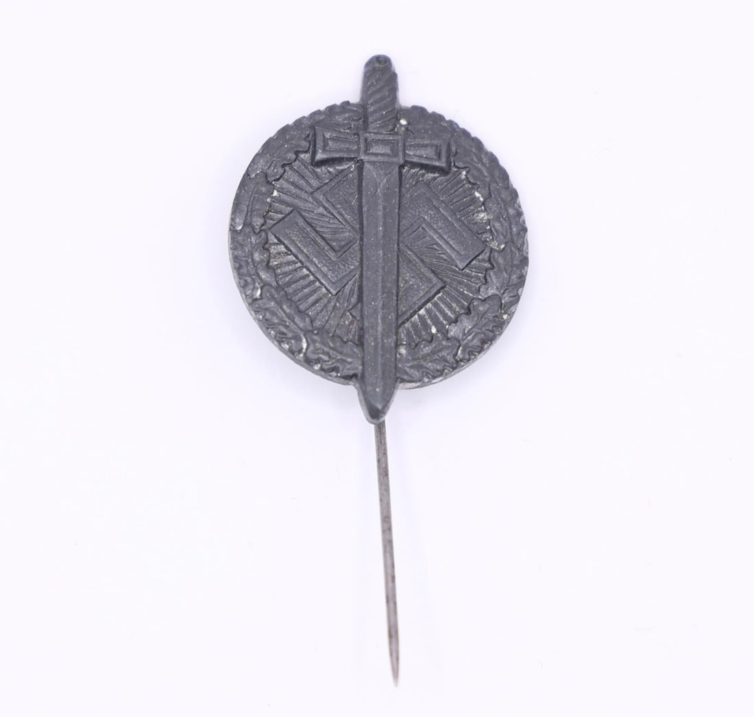 German WWII Tinnie - "Tag der Wehrmacht": A small WWII-era German fundraising tinnie featuring a raised sword across a round wreath with geometric backing. Made of darkened zinc alloy with a vertical pin on the reverse, marked "H4." These wer