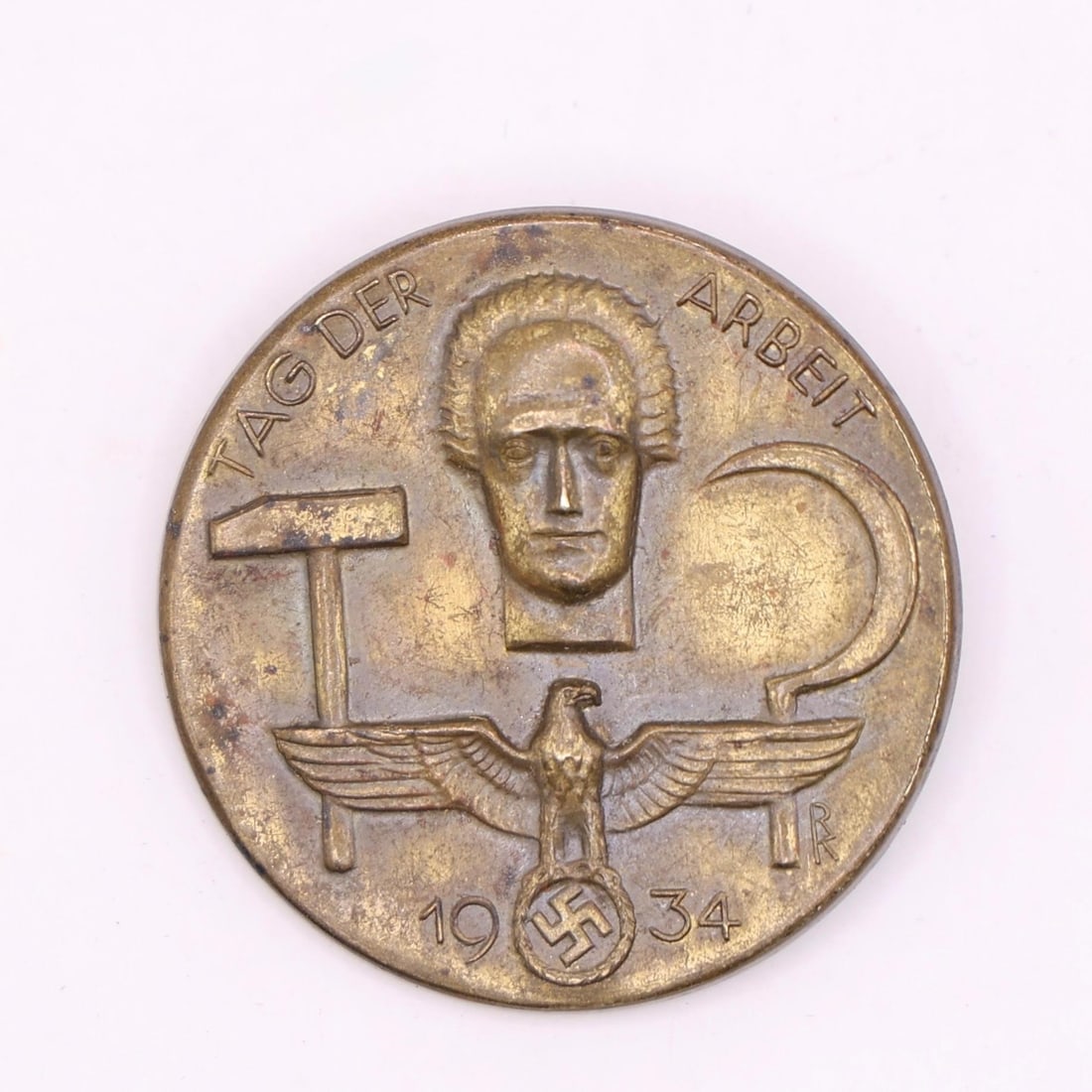 1934 German Labor Day Badge (Bronze Tone): A round 1934 commemorative German Labor Day badge in bronze tone. The front shows a hammer, a sickle, and a stylized face above an eagle emblem. The back has the original pin, though aged. Displays