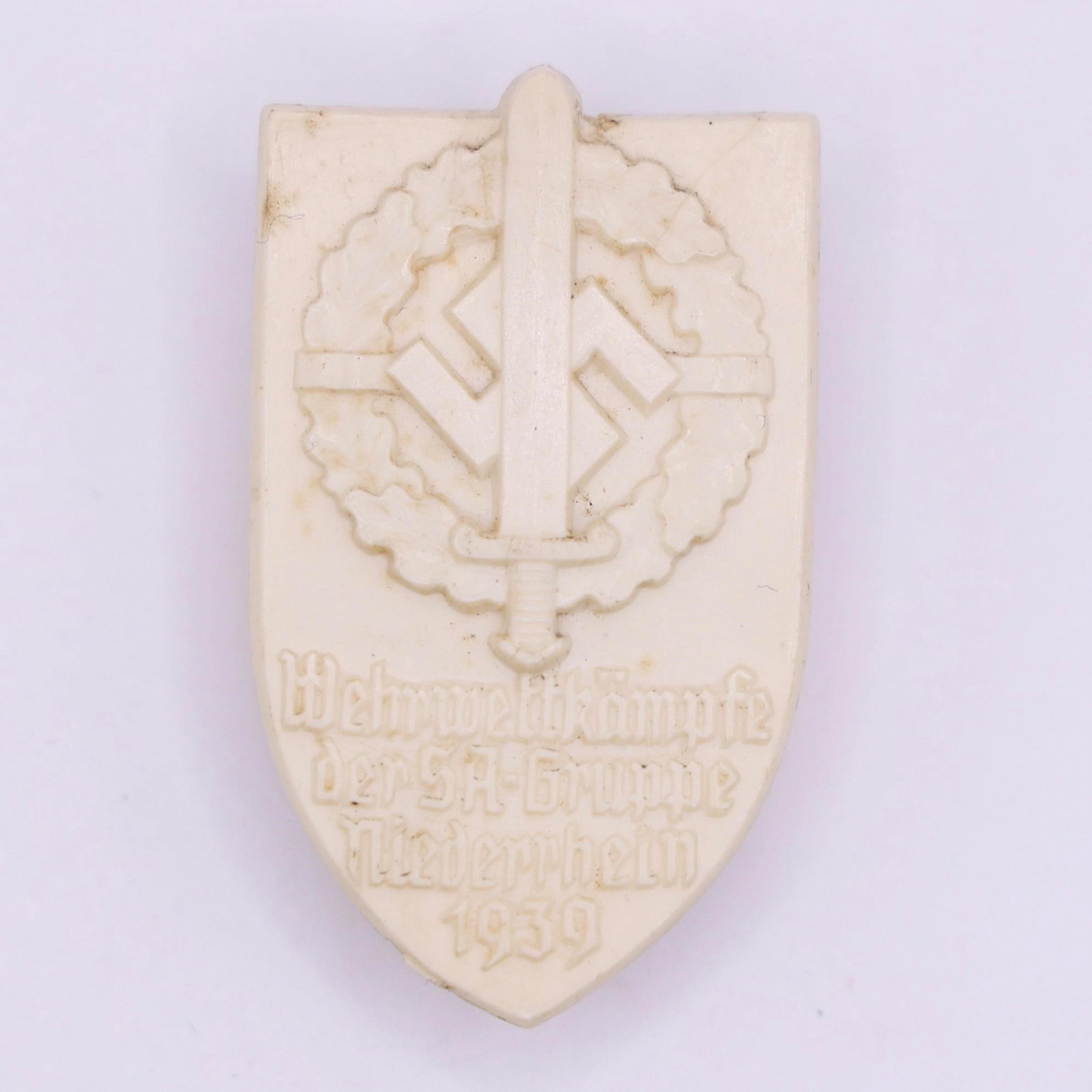 1939 SA Group Niederrhein Sport Day Badge: White protoplastic badge, approximately 35 × 50 mm, originally with bronze-colored paint that has faded over time. Features a sword with SA insignia on the pommel over an oak leaf wreath, with emboss