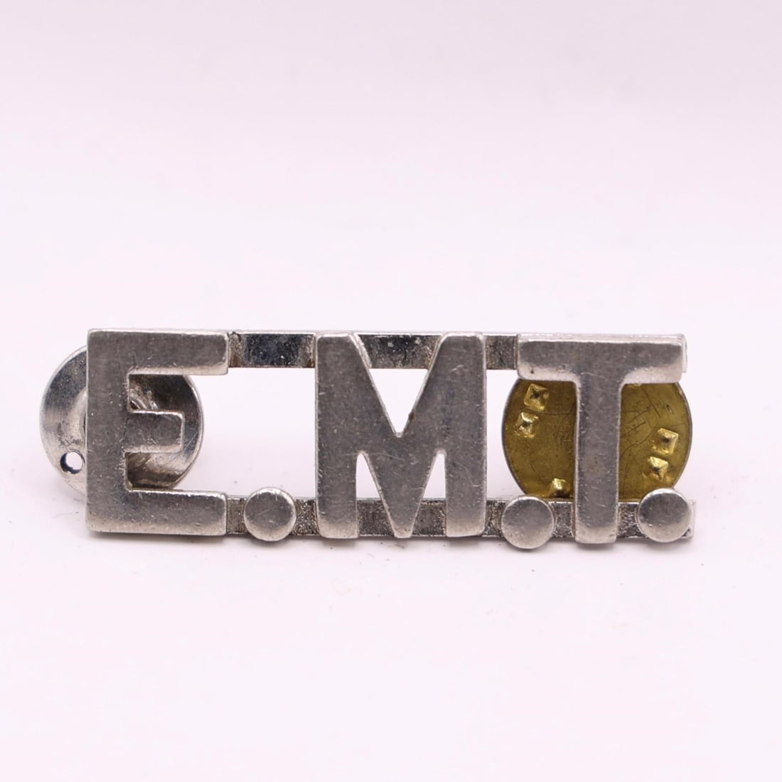 EMT Silver-Tone Metal Collar Insignia Pin: This is an EMT silver-tone metal collar insignia pin. It spells out EMT (Emergency Medical Technician). The pin is typically made of die-struck metal with a silver or nickel finish. It measures