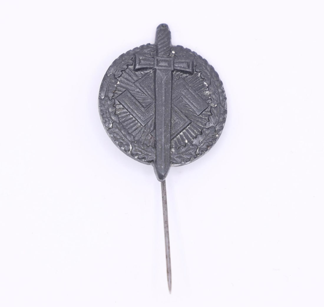 German WWII Tinnie - "Tag der Wehrmacht": A small WWII-era German fundraising tinnie featuring a raised sword across a round wreath with geometric backing. Made of darkened zinc alloy with a vertical pin on the reverse, marked "H4." These
