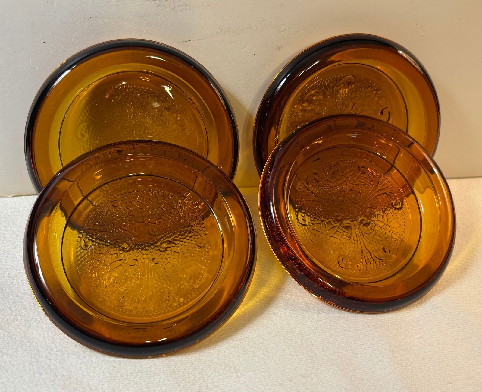 Vintage (1970s) Set of 4 Indiana Glass Tiara Sandwich Amber Glass Coasters: This lot features a set of 4 Indiana glass Tiara sandwich Amber glass coasters. Material: Made of pressed glass featuring a rich amber color. They have an intricate embossed floral and scroll "Sandwic