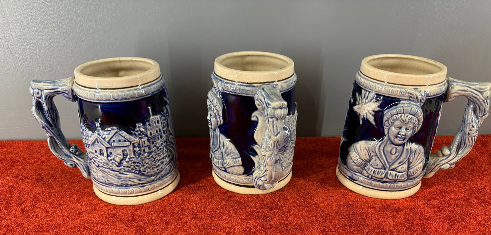 Vtg Trio of (West German) Cobalt Blue Ceramic Beer Steins: Three vintage ceramic beer steins (tankards) featuring high-relief molded scenes on a cobalt blue glazed ground. They mimic traditional German salt-glazed stoneware (Westerwald style), which heavily f