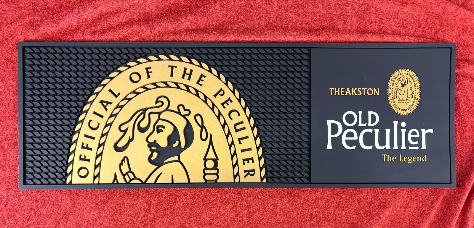 (British) Theakston Old Peculiar Rubber Bar Runner/Mat: Official rubber bar runner mat featuring the branding for Theakston Old Peculier, one of Britain’s most iconic and celebrated traditional strong old ales. The distinctive artwork and wording on the 