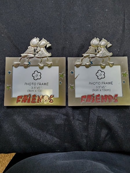 CUTE FRIENDS METAL PICTURE FRAMES: Adorable matching Friends-themed photo frames featuring two cheerful cartoon figures sitting together on top. Each frame is crafted from sturdy metal with colorful star accents and the word “FRIENDS