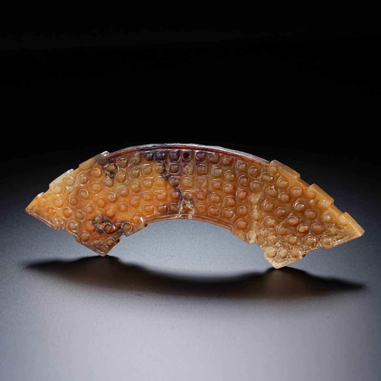 A CHINESE JADE HUANG PENDANT WITH 'DRUM' MOTIF: This jade object is shaped as a flat, arched jade segment, resembling a crescent bridge with both ends diagonally truncated. The outer and inner curved edges feature regularly spaced notches and stepp