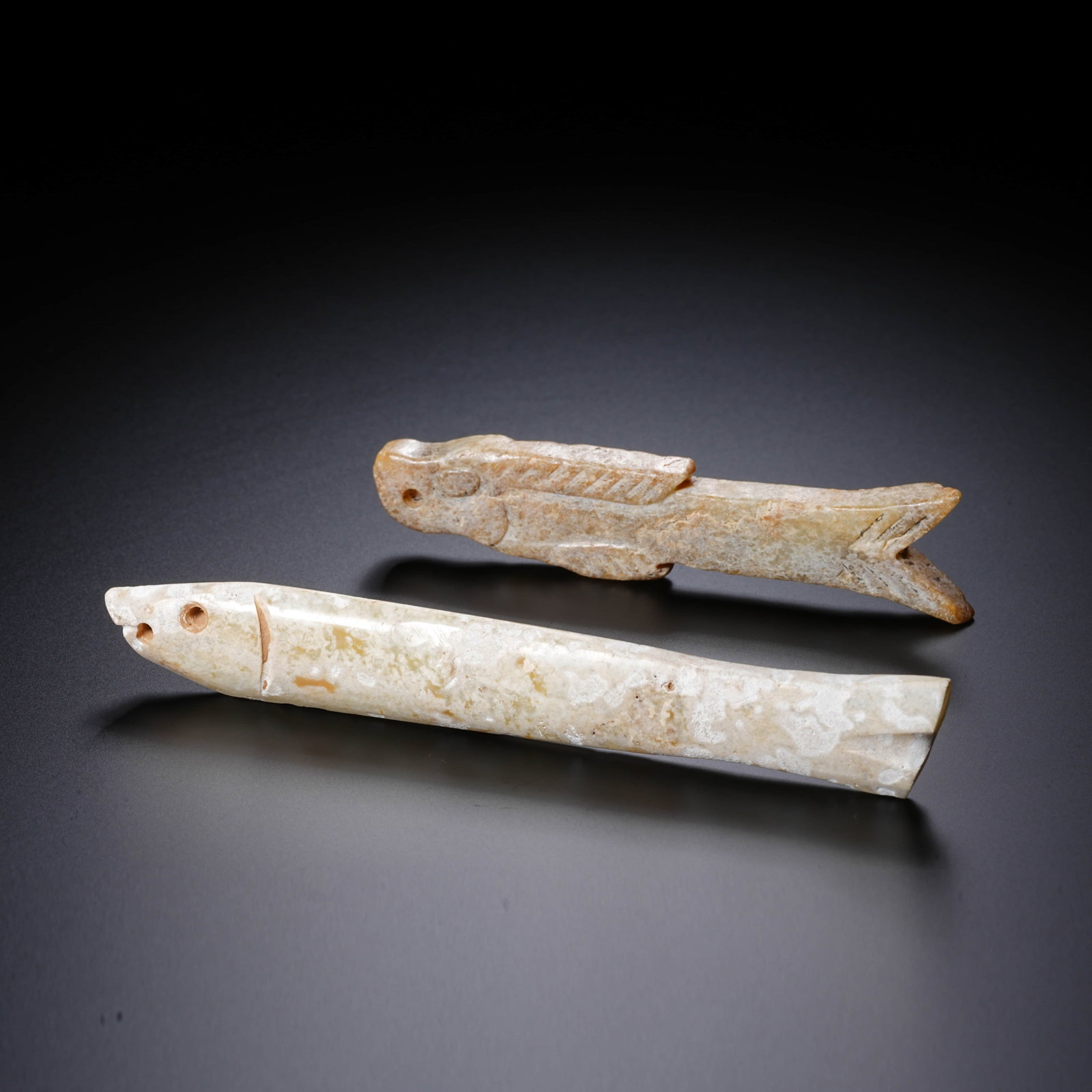 A SET OF CHINESE JADE FISH: This pair consists of two fish-shaped elongated jade ornaments. The fish in the foreground has a relatively straight, cylindrical body with a diagonally truncated mouth drilled with a perforation. The