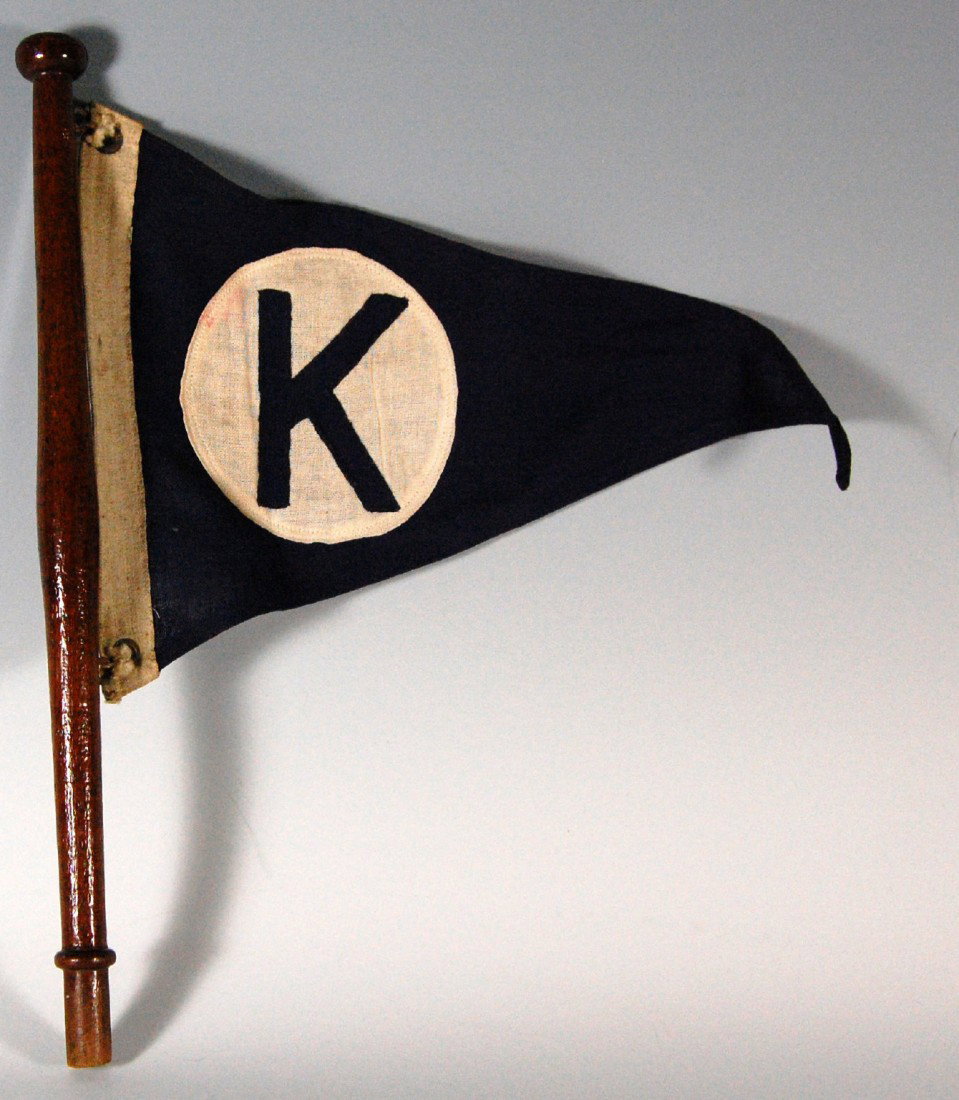 Flag, motor yacht, wool, Initial 'K' Yacht flag, p: Yacht flag, pennant shaped, blue with a white circle and blue "K" within. Constructed of appliqued wool and mounted on what is likely the original turned wood holder and fastened with tied cord throug