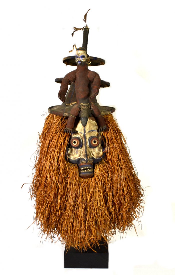 Initiation Mask, Yaka, African: Yaka Tribal Mask Age: Mid 20th Century Materials: Wood Description: The mask is painted with multiple colors and has a raffia surround. On the top rests a three tier hat and a male figure. Size: On St
