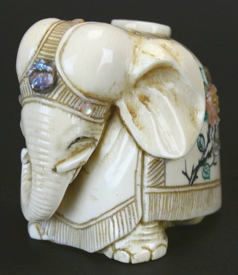 Netsuke, Carved Ivory Ornate Elephant, Signed: Description: A detailed ivory carving of an elephant draped with a highly decorated blanket. Scrimshaw details and branches and leaves. The flowers are mother of pearl and abalone. There's even an aba