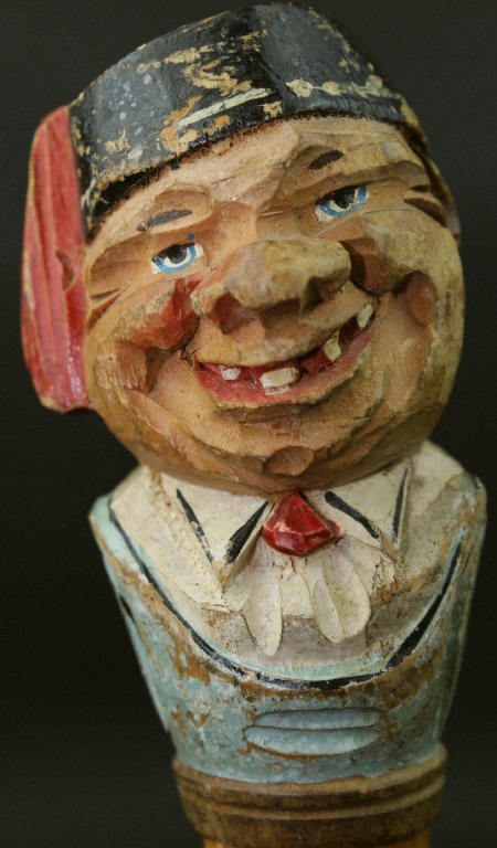 Cane, Beech, Carved Head, C. 1900: This cane or walking stick is made of beech wood with a carved head, likely made of pine, as the grip. The head is that of a man smiling, lips parted to reveal teeth far apart and yellowed. The man is