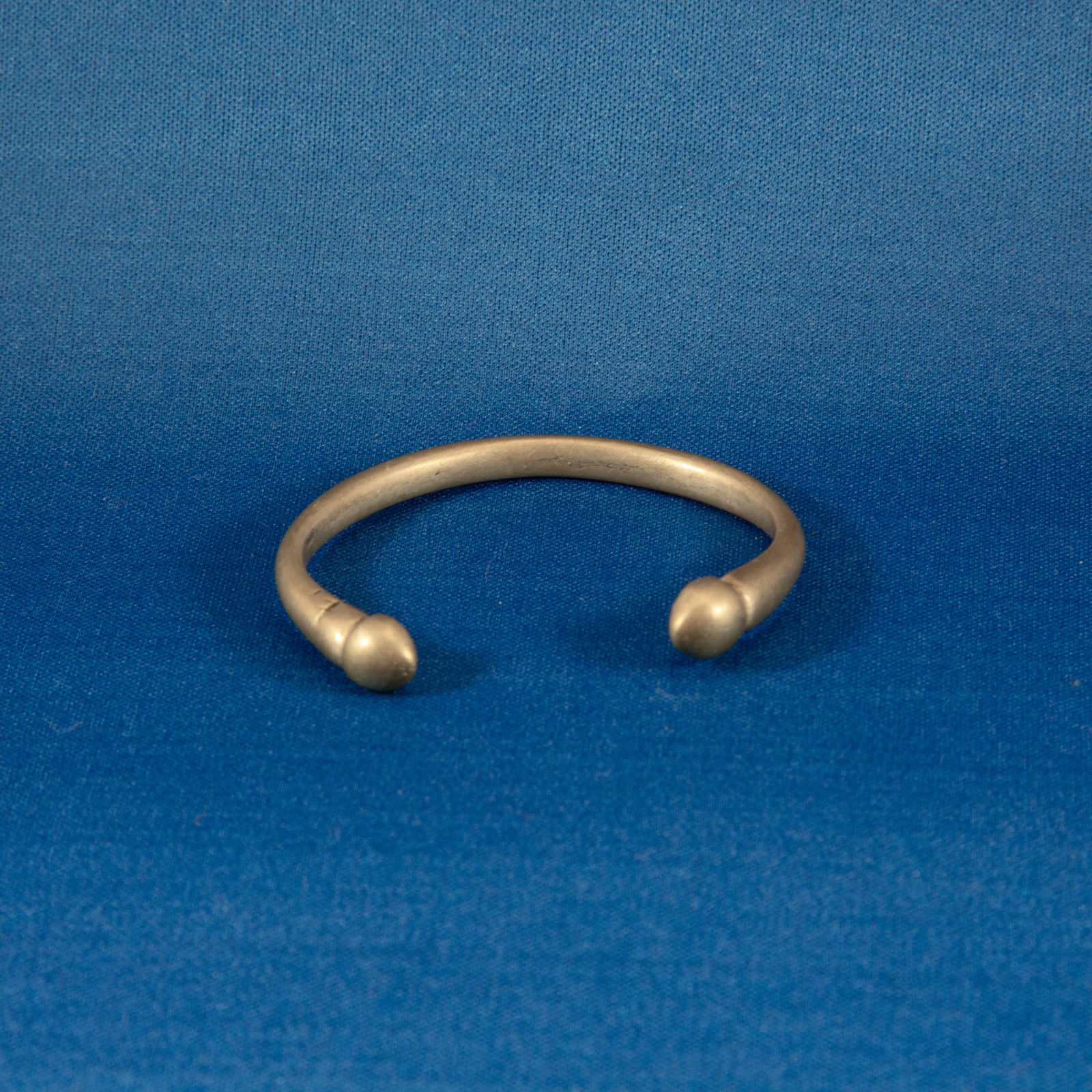 BRACELET: SILVER SASANIAN (224-651 CE): A solid silver Sasanian penannular bracelet dating to the 2nd–7th century AD, formed from a round-section silver rod bent into an open-ended hoop with smoothly rounded, globular terminals. The surfa