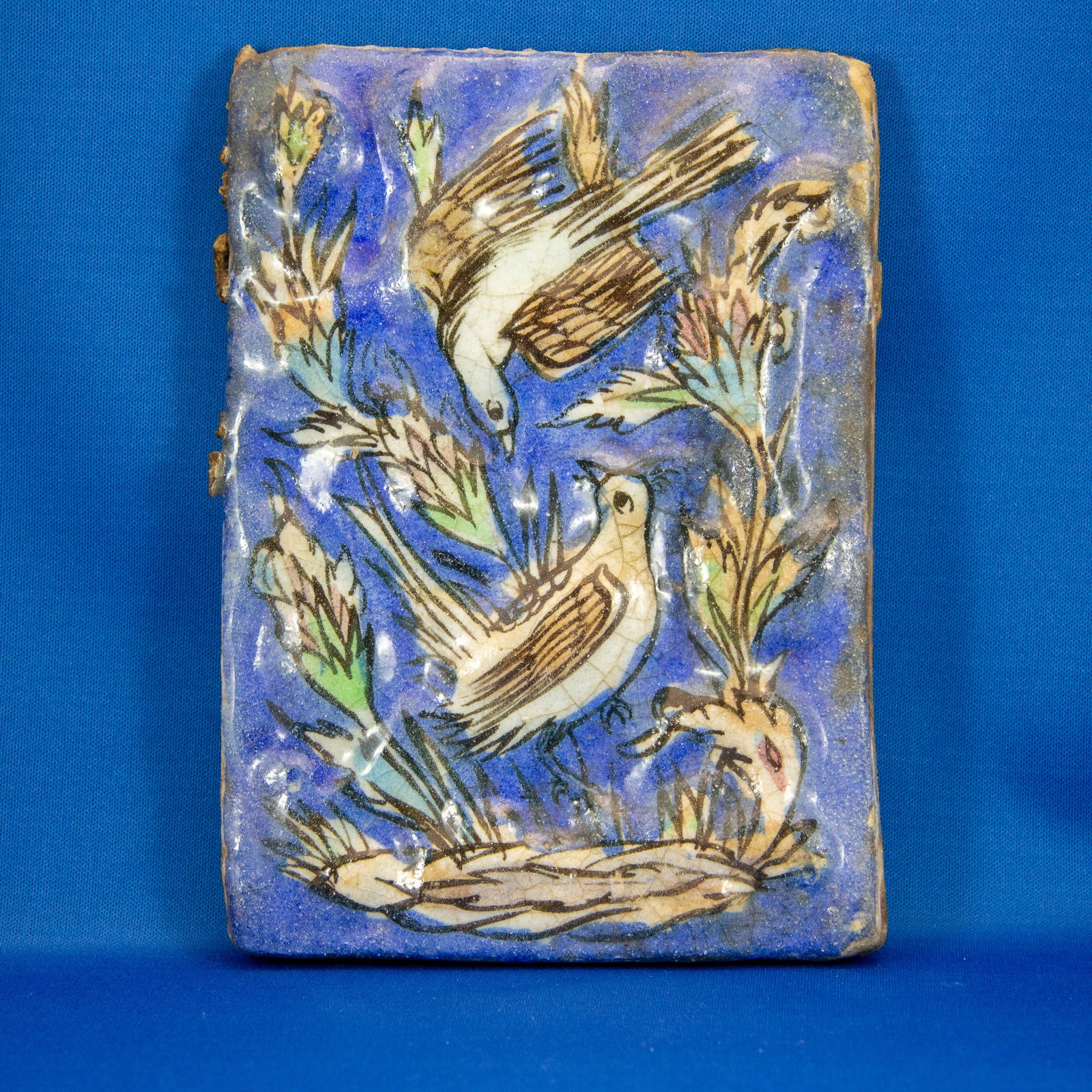 TILE: BIRDS QAJAR (1789-1925 CE): A beautifully painted Qajar-era ceramic tile featuring a lively naturalistic scene of two birds among stylized foliage. The entire composition is set against a rich cobalt-blue ground, a highly desira