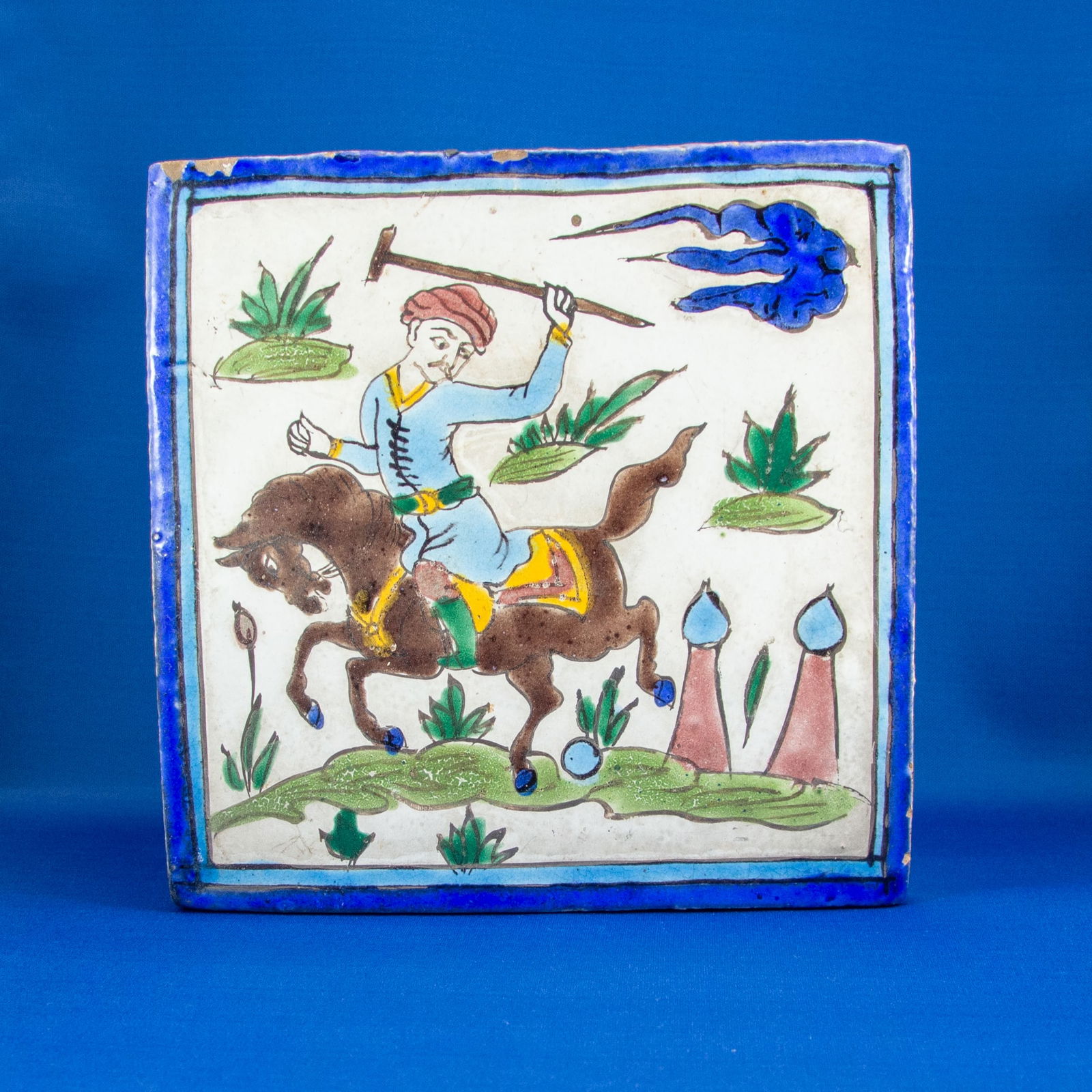 TILE: EQUESTRIAN QAJAR (1789-1925 CE): A vibrant Qajar-era painted ceramic tile depicting an energetic equestrian scene. At the center, a horseman in traditional attire is shown mid-gallop, his body leaning forward with dynamic motion. He