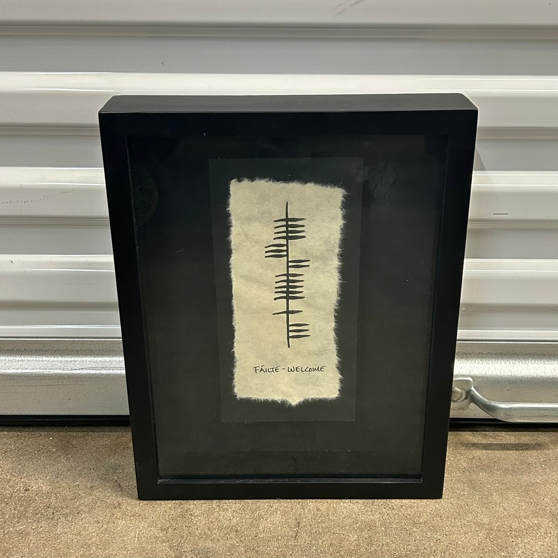 Framed 'Fáilte – Welcome' Handmade Paper Artwork: Framed artwork featuring a minimalist black symbol on deckle-edge handmade paper, inscribed “Fáilte – Welcome.” Black frame with black mat. Approx. 13 in H x 13 in W overall (se