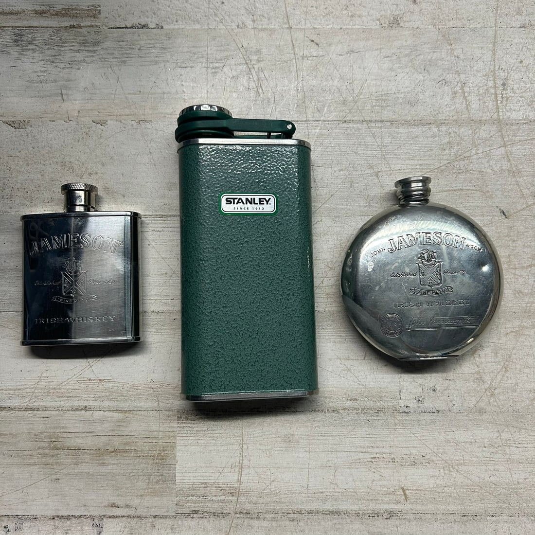 Three Assorted Hip Flasks Including Stanley and Jameson Branded Examples: Group of three metal hip flasks including a green Stanley hammer-tone finish flask marked “Stanley Since 1913,” along with two Jameson Irish Whiskey branded stainless steel flasks. One Jam