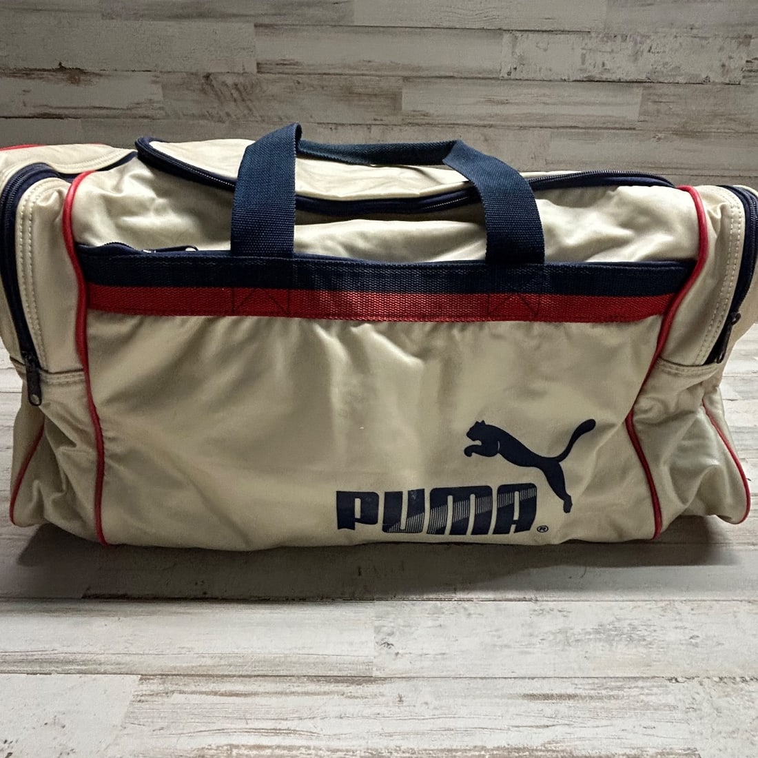Vintage Puma Cream/Navy/Red Striped Duffle Bag Athletic Gym Travel Bag: This vintage Puma duffle bag features a cream-colored, slightly iridescent material accented with navy blue handles and trim, and red and navy stripes. The iconic Puma logo is prominently displayed on