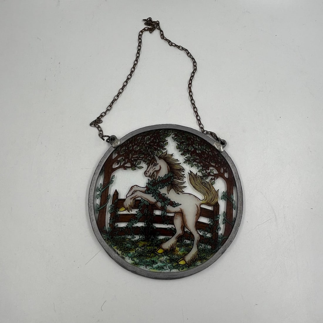 Vintage Stained Glass Suncatcher Horse in Paddock Art Decor 1988 Mark: This charming vintage stained glass suncatcher depicts a graceful white horse in a paddock, with a wooden fence and lush greenery. The round piece is framed in metal and hung with a chain for display