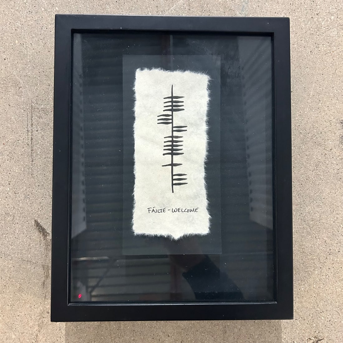 Framed Ogham Inscription 'Fáilte - Welcome' Art Piece on Textured Paper: This unique framed art piece features a striking Ogham inscription, a historic form of writing, translating to 'Fáilte - Welcome'. The inscription is rendered in bold black ink on a textured, handmad