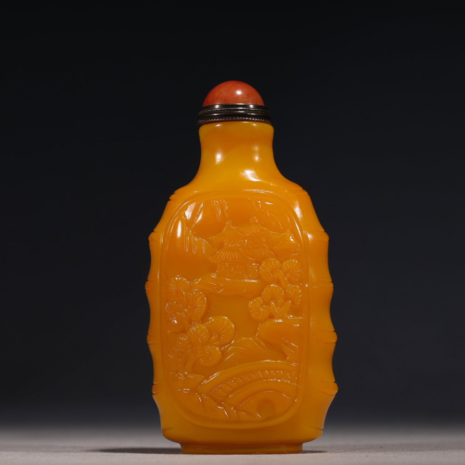 A Chinese Yellow Glass Snuff Bottle Carved with Landscape,Qianlong Mark: A Chinese opaque yellow glass snuff bottle of flattened ovoid form,carved in relief with a pavilion,trees,and rocks within a shaped panel.It is fitted with a red stone stopper.The base bears a four-ch
